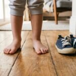 Kind steht barfuß auf einem Holzboden neben einem Paar Kinderschuhe